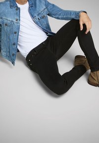 Chaqueta de mezclilla en azul claro, camiseta blanca ajustada, jeans negros ajustados y botas de tobillo color beige. Texturas suaves, botones simples y diseño a medida.