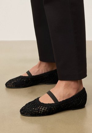 Sorte hæklede balletsko med elastikrem båret med ankel-lange sorte bukser på en beige gulv.