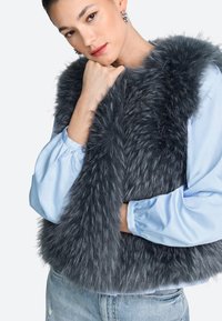 Grijze nepbontvest met een fluwelige textuur. Draagt over een lichtblouse met pofmouwen. Verpauperde blauwe spijkerbroek maakt de outfit compleet.