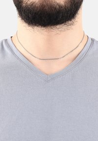 Collana in argento indossata sopra una maglietta grigia a scollo a V, caratterizzata da piccoli anelli e una superficie liscia. La maglietta ha una finitura a coste vicino al collo.