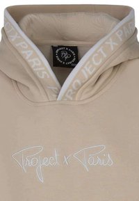 Sweat à capuche beige avec un logo brodé blanc "Project x Paris" sur la poitrine, présentant une texture douce et un col intérieur blanc contrastant.