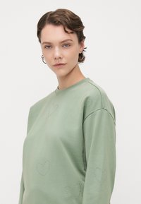 Grønn sweatshirt i myk stoff, med dekorative hjerteformede nagler på forsiden. Rund hals, avslappet passform og lange ermer.