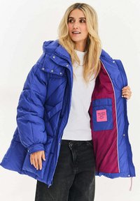 Modrá prošívaná péřová bunda s kapucí, vybavená cvočky, elastickými manžetami a vnitřní kapsou s růžovou podšívkou. Viditelné kontrastní prošívání.