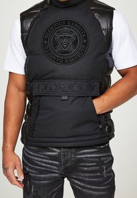 Gilet matelassé noir avec un logo rond embossé, une fermeture éclair et des sangles latérales réglables. Poche avant avec des accents en tissu texturé.