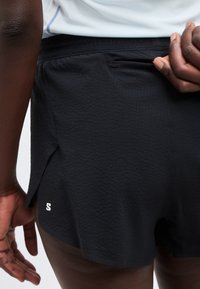 Κοντινό πλάνο ατόμου που φοράει μαύρα αθλητικά σορτς με υφή και μικρή τσέπη με φερμουάρ στο πίσω μέρος, τραβώντας τη μέση με το ένα χέρι.