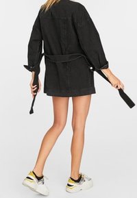 Person, der eine übergroße schwarze Jeansjacke mit einem Gürtel und weiße Sneakers mit gelben und schwarzen Details trägt, steht vor einem schlichten Hintergrund.