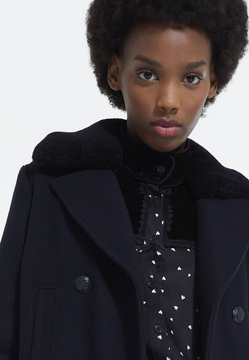 Femme avec des cheveux courts et bouclés portant un manteau sombre avec un col en fourrure sur une chemise noire à motifs de cœurs blancs.