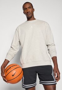 Felpa grigia chiara con collo a giro e logo bianco, abbinata a pantaloni corti neri perforati con strisce bianche sui lati. Tiene in mano un pallone da basket.