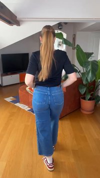 Hosszú lófarokkal rendelkező nő sétál a nappaliban fekete ingben, kék farmerban és piros-fehér sportcipőben.