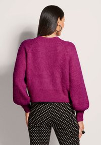 Fuchsia gestrickter Pullover mit puffigen Ärmeln und geripptem Saum, kombiniert mit schwarzen Hosen mit einem weißen quadratischen Muster.