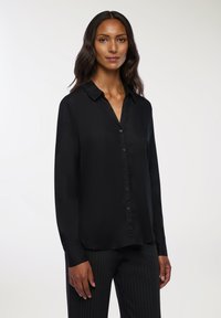 Czarna bluzka z długim rękawem i kołnierzem, z przodem zapinanym na guziki i gładką, błyszczącą teksturą. Noszona z spodniami w prążki.
