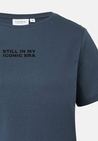 Niebieski bawełnowy T-shirt z krótkimi rękawami i okrągłym dekoltem. Z przodu widoczny czarny napis "STILL IN MY ICONIC ERA". Gładka tekstura. Widoczna metka rozmiaru.