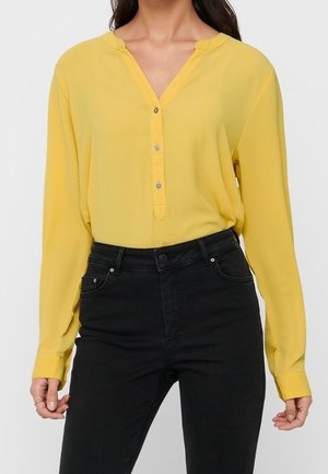 Nainen, jolla on keltainen pitkähihaisen napitettava paita ja mustat korkeavyötäröiset farkut, seisoo käsivarret rentoina sivuilla.