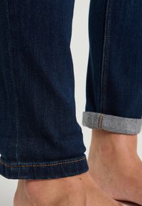 Jeans de mezclilla azul oscuro con puño gris claro. Cuentan con costuras naranjas y un diseño ajustado. El material tiene una textura suave.