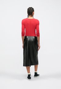 Persoon met ingevlochten haar, gekleed in een rood shirt met lange mouwen, een zwarte leren midirok, witte sokken en zwarte hoge sneakers, van achteren gezien.