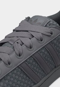 Κοντινό πλάνο γκρι αθλητικού παπουτσιού με υφή ύφανσης, χοντρές κορδόνια, ραμμένες ρίγες και λογότυπο στη μαξιλαρωμένη γλώσσα.