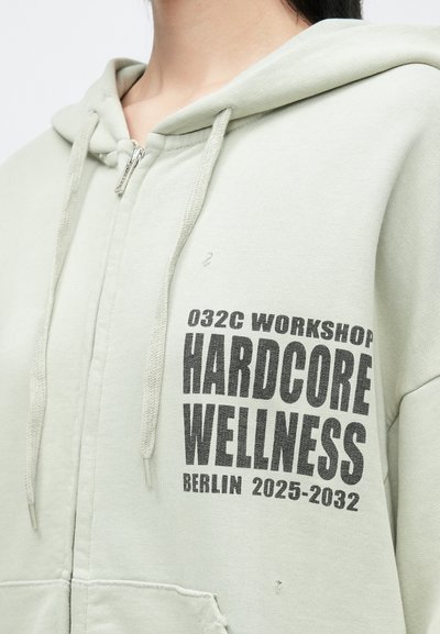 Világos zöld cipzáros kapucnis pulcsi, megviselt textúrával, fekete nyomtatott szöveggel: "032C WORKSHOP HARDCORE WELLNESS BERLIN 2025-2032."