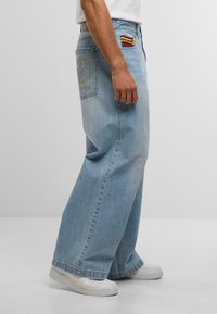 Mann in hellblauen Schlagjeans mit bestickter Gesäßtasche und mehrfarbigem Stoffdetail in der Taille, kombiniert mit weißen Turnschuhen.