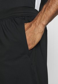 Mustat urheiluhousut, joissa on sileä pinta, joustava vyötärönauha ja sivutasku, johon käsi on asetettu.