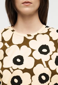 Γυναίκα που φοράει μια μπεζ πουκάμισο με μεγάλα λευκά λουλούδια που έχουν μαύρα κέντρα, ορατή από τον λαιμό μέχρι το πάνω μέρος του στήθους, σε απλό φόντο.