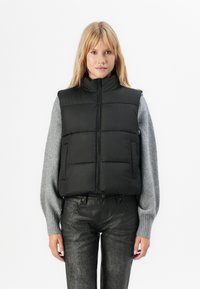 Vrouw in een zwarte gewatteerde vest over een lange mouw grijze trui en zwarte gestructureerde broek, staand tegen een witte achtergrond.