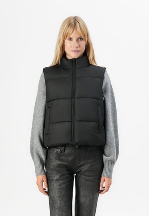 Vrouw in een zwarte gewatteerde vest over een lange mouw grijze trui en zwarte gestructureerde broek, staand tegen een witte achtergrond.