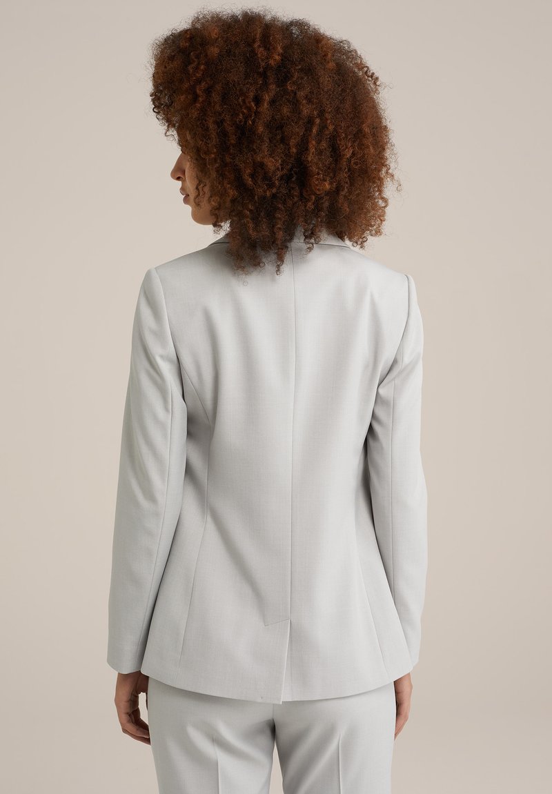 Blazer ajusté gris clair avec des épaules structurées, une fente au dos et un tissu lisse. Porté sur un pantalon assorti, arrière-plan uni.