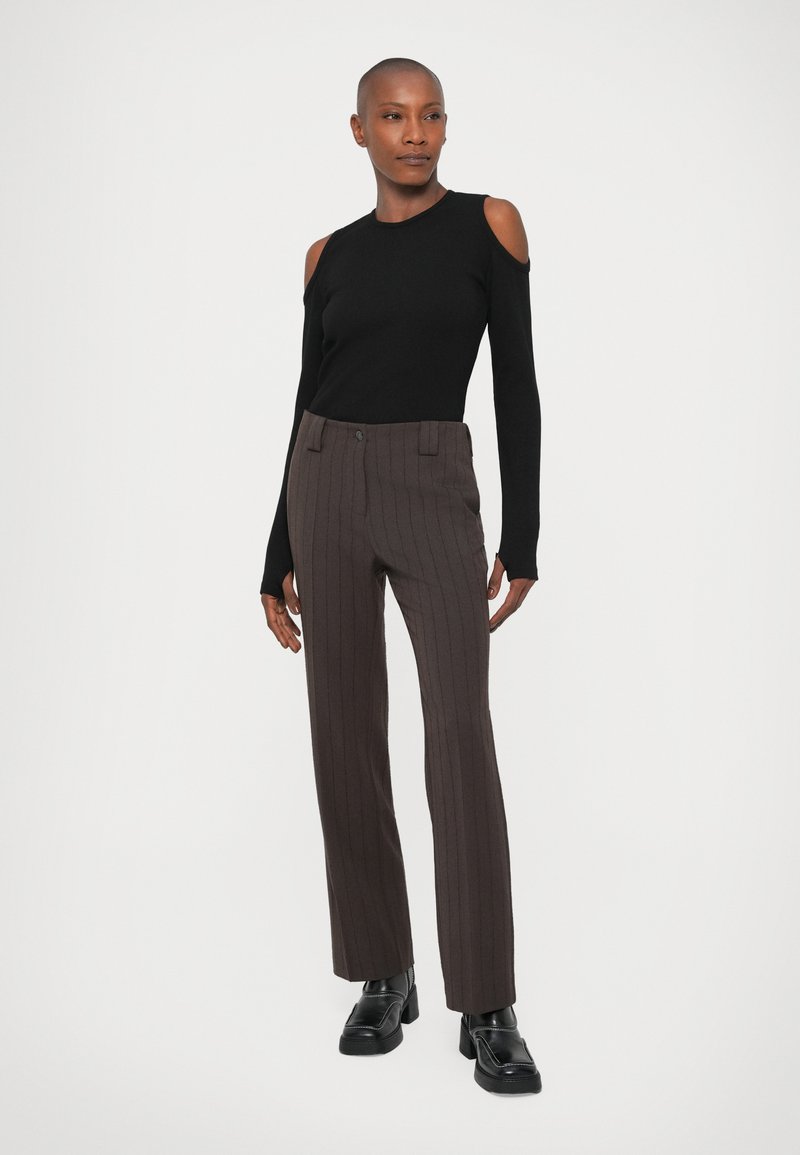 Haut noir à épaules nues et pantalon marron à rayures, offrant une coupe ajustée. Des chaussures noires épaisses complètent la tenue sur un fond uni.