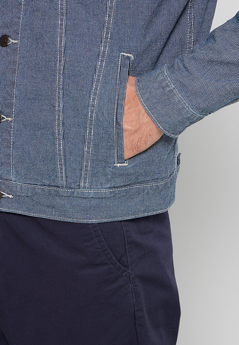 Veste en denim avec des rayures verticales, couleur bleu foncé, boutons en métal et poches latérales. Les poignets et l'ourlet sont légèrement contrastés.