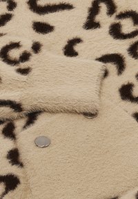 Bársonyos, bézs színű szövet barna leopárd pöttyökkel, texturált felülettel és fényes patentgombokkal. Részletei között egy kis zseb található a szélén.