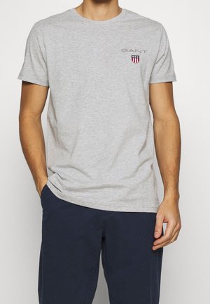 Mann trägt hellgraues kurzärmeliges T-Shirt mit kleinem GANT-Logo und marineblaue Hose, eine Hand in der Tasche, die andere entspannt.