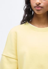 Ανοιχτό κίτρινο φούτερ με ριγωτό λαιμόκοψη, φαρδιά μανίκια και απαλό υλικό, εμφανίζεται μερικώς πάνω σε απλό λευκό φόντο.