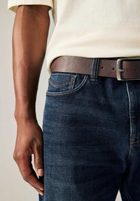 Dark blue denim jeans with a textured finish, accented by orange stitching. A brown leather belt with a silver metal buckle is visible.