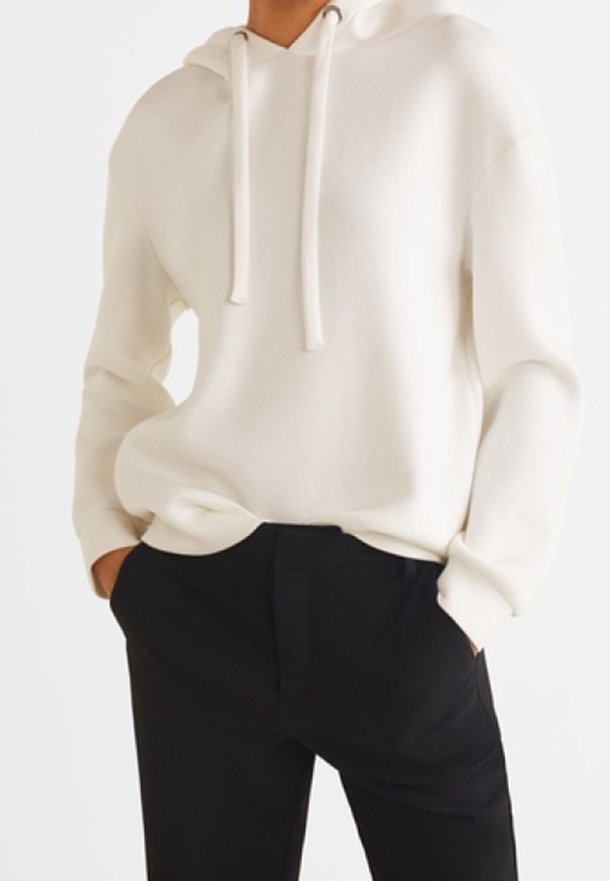 Sudadera crema con un corte relajado y cordones ajustables. Combinada con pantalones negros, que muestran una textura suave y un diseño minimalista.
