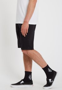 Shorts en jean noir, t-shirt en coton blanc, chaussures noires sans lacets avec des accents blancs, et chaussettes noires avec un logo.