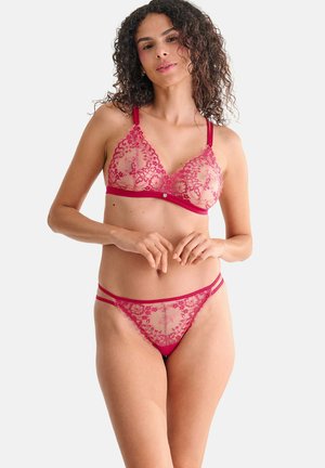 Femme aux cheveux bouclés portant un soutien-gorge en dentelle rouge et une culotte en dentelle assortie sur un fond clair uni.