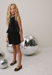 Crni baršunasti top s peplum detaljem i kratke hlače, upareni s ravnim cipelama s leopardovim uzorkom. U pozadini sjaje disko kugle na svijetlom podu.