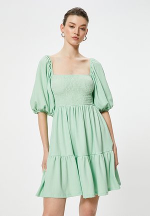 Vestido verde menta con escote cuadrado, mangas abullonadas y volantes en capas en el dobladillo. La textura parece ligera y ligeramente arrugada.