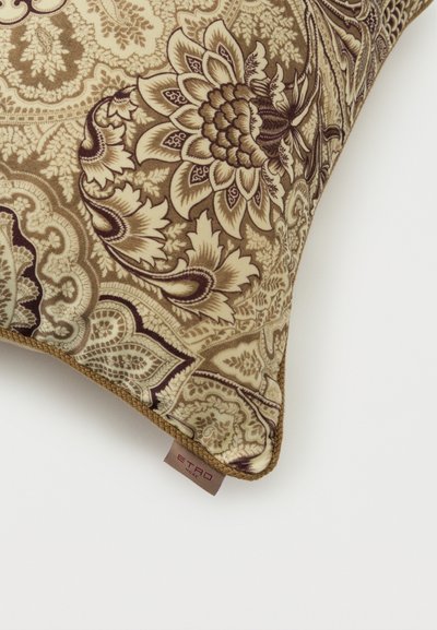 Coussin avec un motif floral cachemire beige et marron complexe et une bordure tressée, affichant une petite étiquette en tissu au bord inférieur.