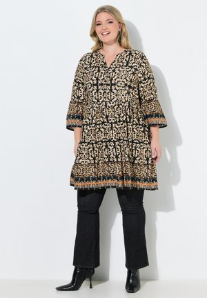 Smilende kvinde iført en mønstret tunika med 3/4 ærmer, sorte bukser og sorte ankelstøvler med hæl, stående mod en hvid baggrund.