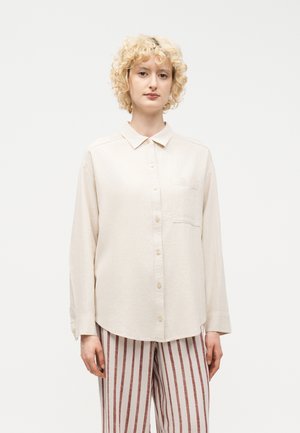 Personne aux cheveux blonds bouclés portant une chemise beige boutonnée et un pantalon taille haute à rayures verticales marron et beige, debout devant un fond blanc.