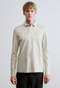 Μακρυμάνικη πόλο μπλούζα σε ανοιχτό μπεζ χρώμα, κατασκευασμένη από απαλό ύφασμα. Διαθέτει κολάρο με κουμπιά και κεντημένο λογότυπο στο στήθος.