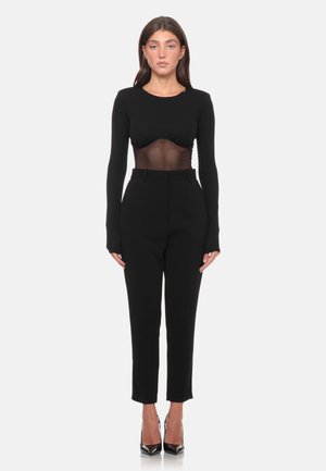 Donna che indossa una maglia nera a maniche lunghe con sezione centrale trasparente, pantaloni neri a vita alta e tacchi neri a punta, in piedi su sfondo bianco.