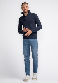 Granatowy sweter dzianinowy z zapięciem przy wysokim kołnierzu, ryflowanym dołem i mankietami, zestawiony z jasnoniebieskimi dżinsami i białymi trampkami, stojący na neutralnym tle.