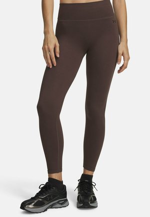 Persoon draagt bruine, nauwsluitende leggings en zwarte sportschoenen, staand tegen een effen witte achtergrond, torso en benen zichtbaar.