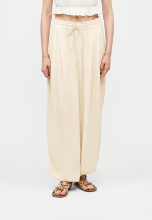 Kobieta w luźnych beżowych spodniach z wiązaniem w pasie, białym krótki topie i brązowych sandałach na paski, stojąca na jednolitym tle.