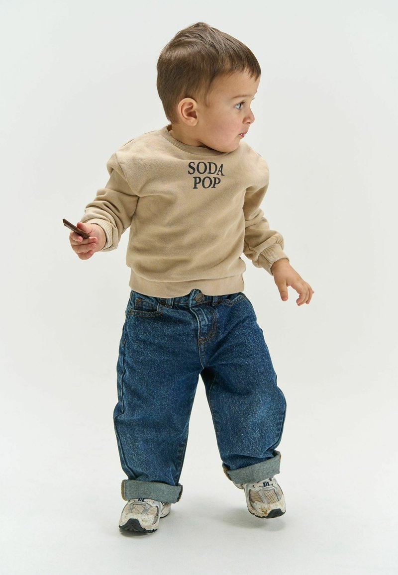 Tout-petit portant un sweat-shirt beige et un jean bleu, tenant un petit objet, regardant vers la droite sur un fond uni.