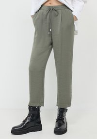 Pantalons légers olive avec une finition texturée, dotés d'une taille élastique avec cordon de serrage et de poches latérales, associés à des bottes de combat noires.