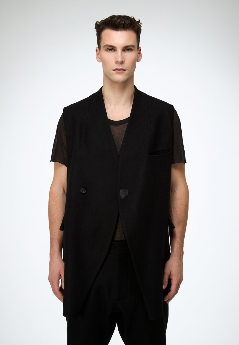 Schwarze, ärmellose Weste mit tailliertem Schnitt. Verfügt über zwei Knöpfe, eine Brusttasche und scharfe Revers. Getragen über einem transparenten schwarzen Oberteil.