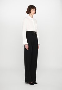 Camicia bianca con colletto e maniche lunghe, abbinata a pantaloni neri a vita alta con doppia fibbia e gambe ampie. Tacchi a punta neri.